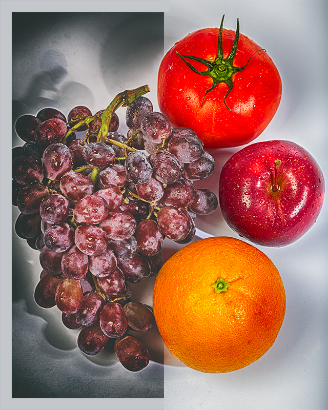 A bushel of grapes, tomato, apple, and orange that are covered in water droplets. They are sitting on a bicolored grungy background.