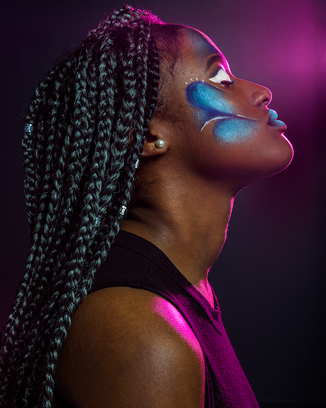 Model Tyra Young poses for a profile shot facing left with her eyes closed. She is wearing a full face of blue, white, and teal makeup that swirls and curves.  Magenta light outlines the edges of her face.