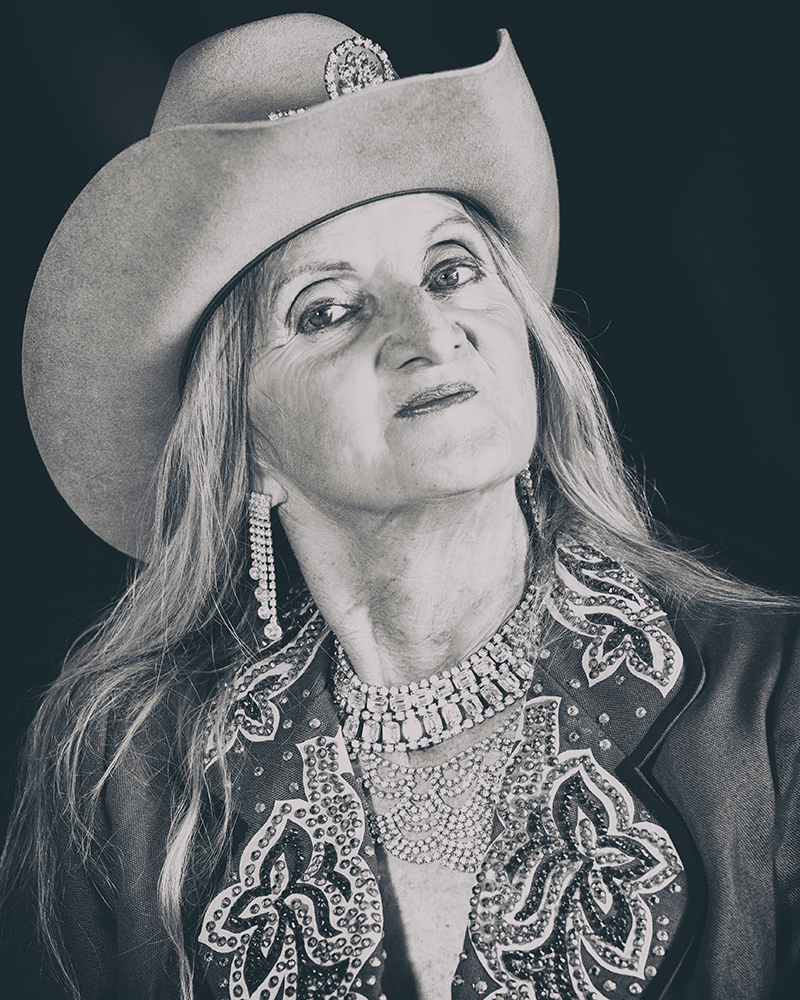 Western rider Patricia Richman poses at a 3/4th angle. Her chin is tilted upward slightly and she's looking confidently toward the camera. Her showcoat has an elegant, sparkling pattern and is matched with earrings and necklace. Pat's cowgirl hat is tilted up, barely revealing the blink around the brim.