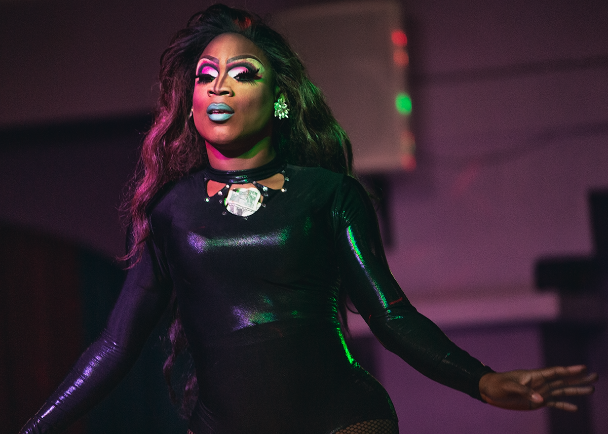 Drag Queen Free Dupree looks on toward the crowd, arms outstretched as she turns gracefully. Her hair flows elegantly and catches in the magenta light of the bar. 