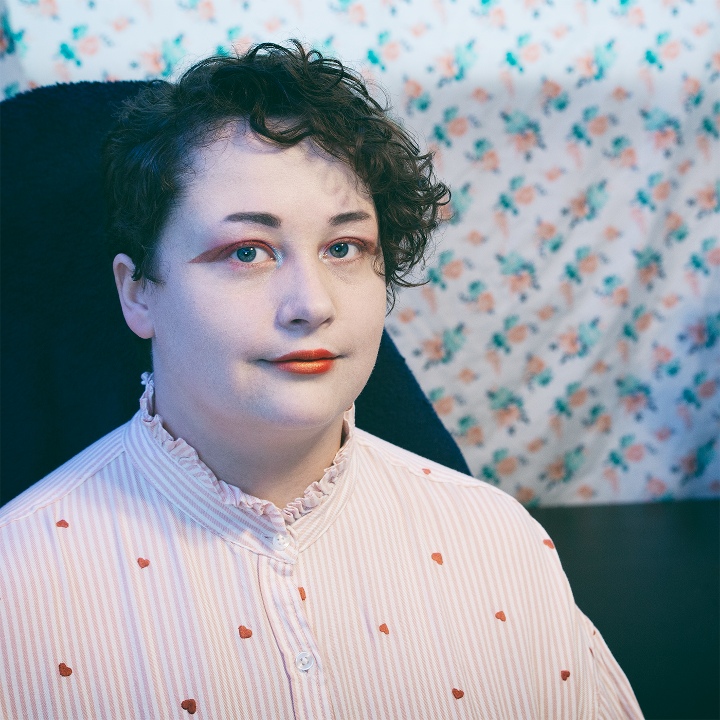 Alejandro sits at his desk in 3/4th view, grey-blue eyes looking up toward the camera. He is wearing a light pink and white stirped button-up with tiny embroidered hearts patterned onto the cloth. He is wearing vibrabt puppy liner that matches his red and gold painted lips. 