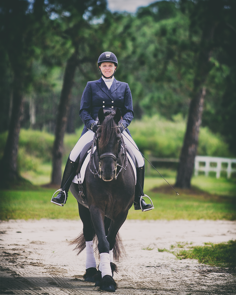 A woman in full show clothes rides down a gravel pathway on a Friesian horse. She is riding toward the camera and smiling. The horse's ears are perking forward and toward the camera. 