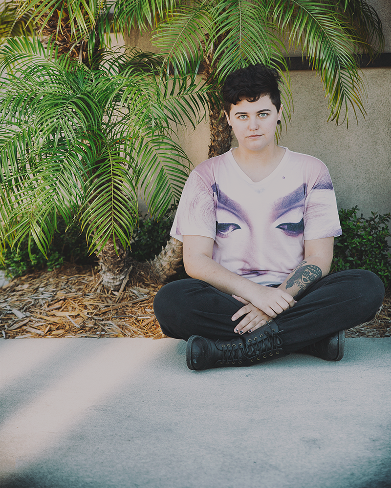 Spencer sits cross legged wearing a graphic t-shirt, black pants, and black boots. He's looking toward the camera with a neutral expression. Behind him are palm trees bathed in light. Two streams of light jut out beside him diagonally on the cement sidewalk he's sitting on.